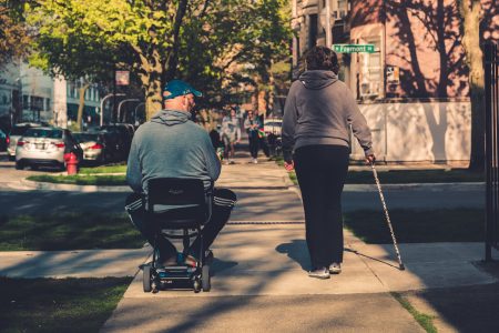 A person in a wheelchair and a person using a cane walking side by side on the sidewalk.