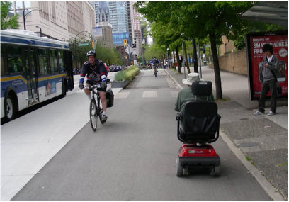 Image of a mobility scooter user and a cyclist passing by each other in a protected cycle lane.  There is a sidewalk to the right and a bus to the left.